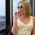 Woman vaping on train in summer attire, sunglasses, bob haircut, enjoying travel, relaxed commute, modern lifestyle.