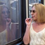 Woman in sunglasses and white top vaping on a train, reflected in window.