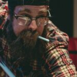 Man with beard and glasses playing pool, wearing a plaid shirt and cap, focused expression, pool chalk in mouth.
