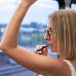Woman with sunglasses applying lip balm on a train, stylish and relaxed travel setting.