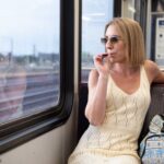 Woman in sunglasses vaping on a train, wearing a white dress, with a patterned backpack, looking out the window.