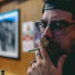 Bearded man with glasses and cap smoking indoors, contemplating, wooden wall background, hipster style.