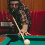 Man playing pool in a bar, focusing on a shot with a cue stick, wearing a plaid shirt and cap, showcasing billiards skills.