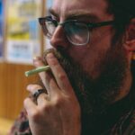 Man with beard and glasses smoking in indoor setting, wearing plaid shirt and cap, relaxed atmosphere.